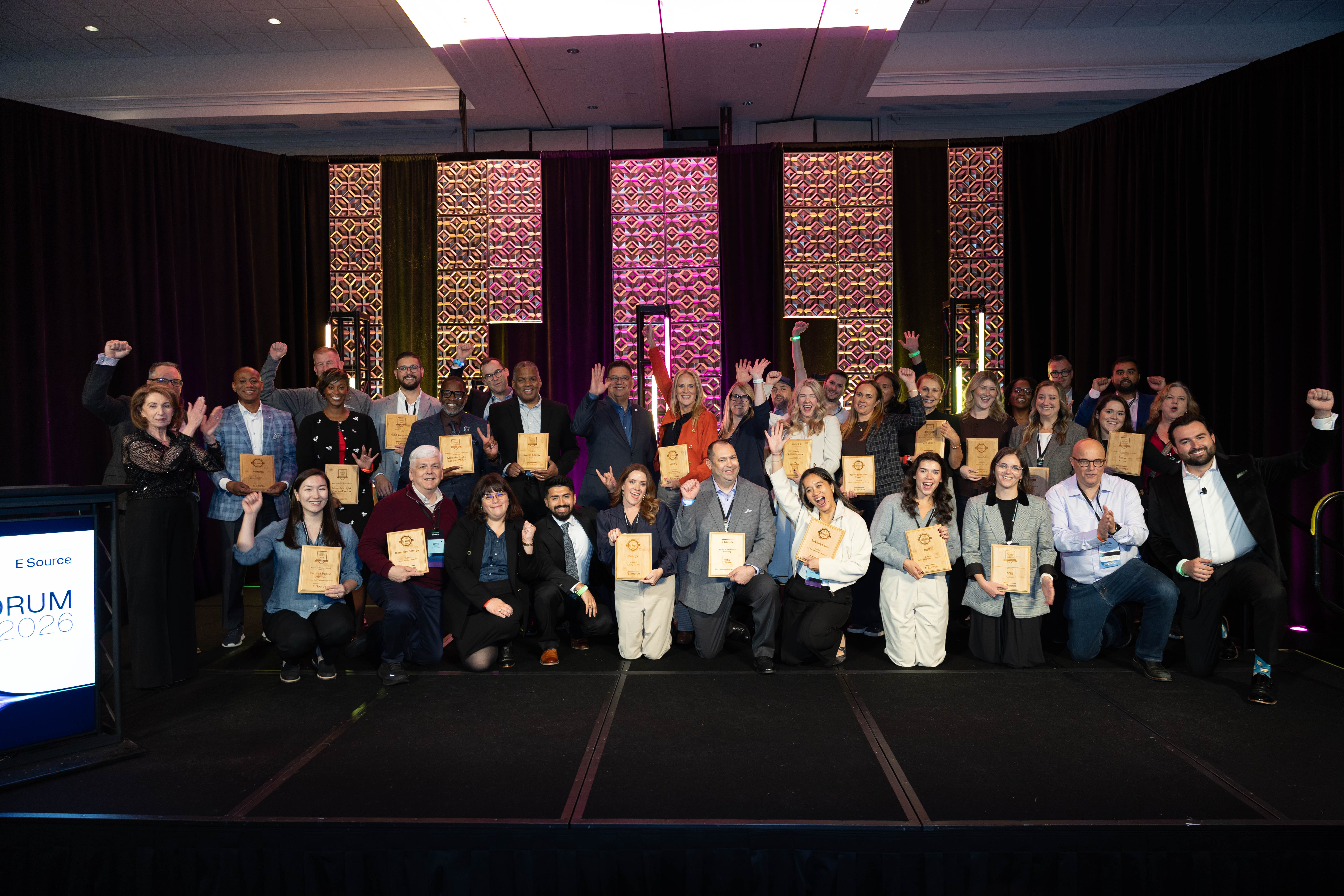 Award winners pose on a stage, many holding wooden plaques and some raising their fists in celebration. Decorative panels lit in pink and purple line the back wall, and a podium on the left displays the E Source Forum 2026 logo.