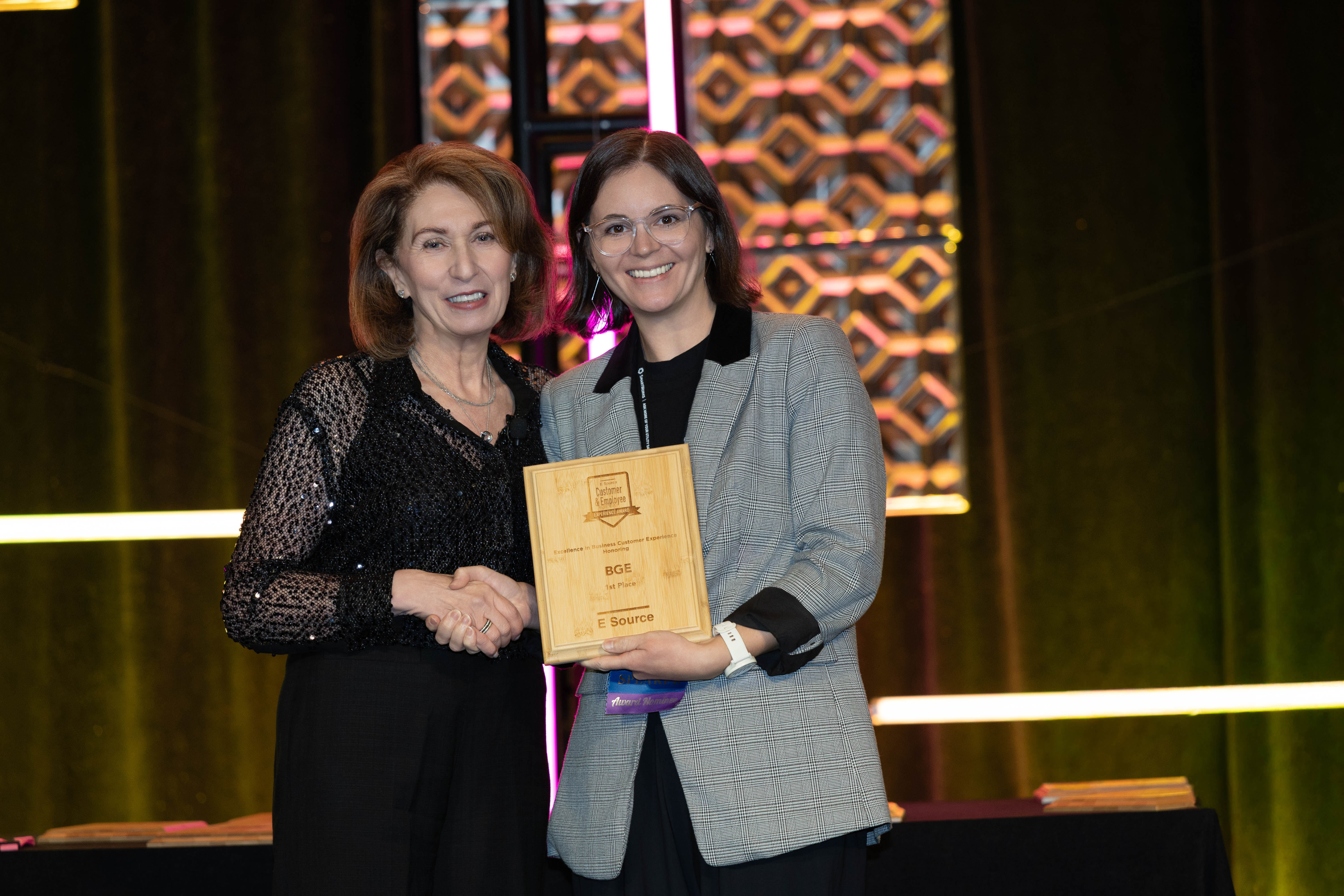 Two smiling women shake hands on stage. One, in a black sequined top, stands beside the other, who wears glasses and a gray plaid blazer and holds a wooden E Source award plaque reading BGE, 1st Place. Pink-lit decorative panels glow behind them.