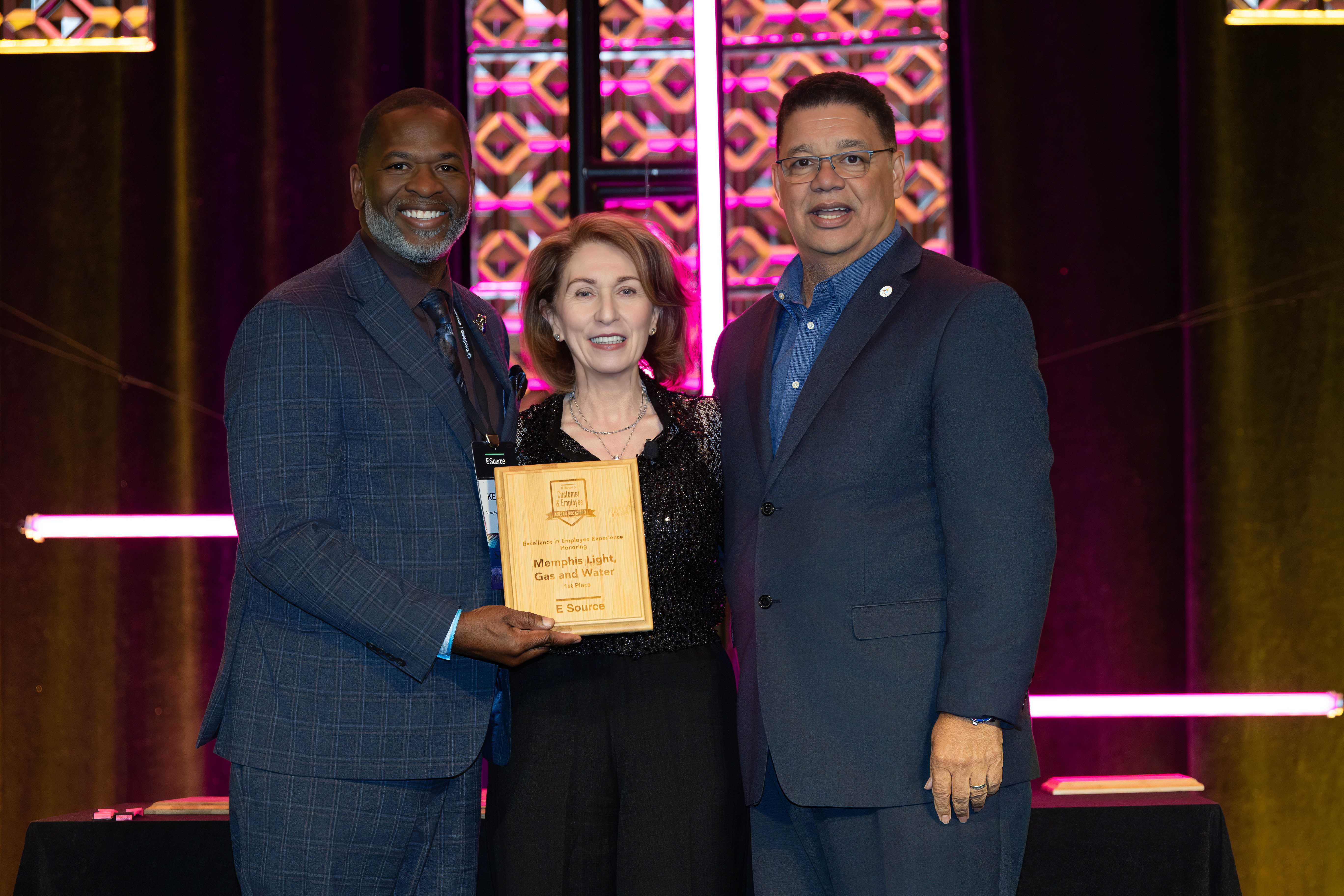 Two men in dark suits stand on stage with a smiling woman in a black sequined top between them. One man holds a wooden E Source award plaque reading Memphis Light, Gas and Water, 1st Place. Pink-lit decorative panels glow behind them.