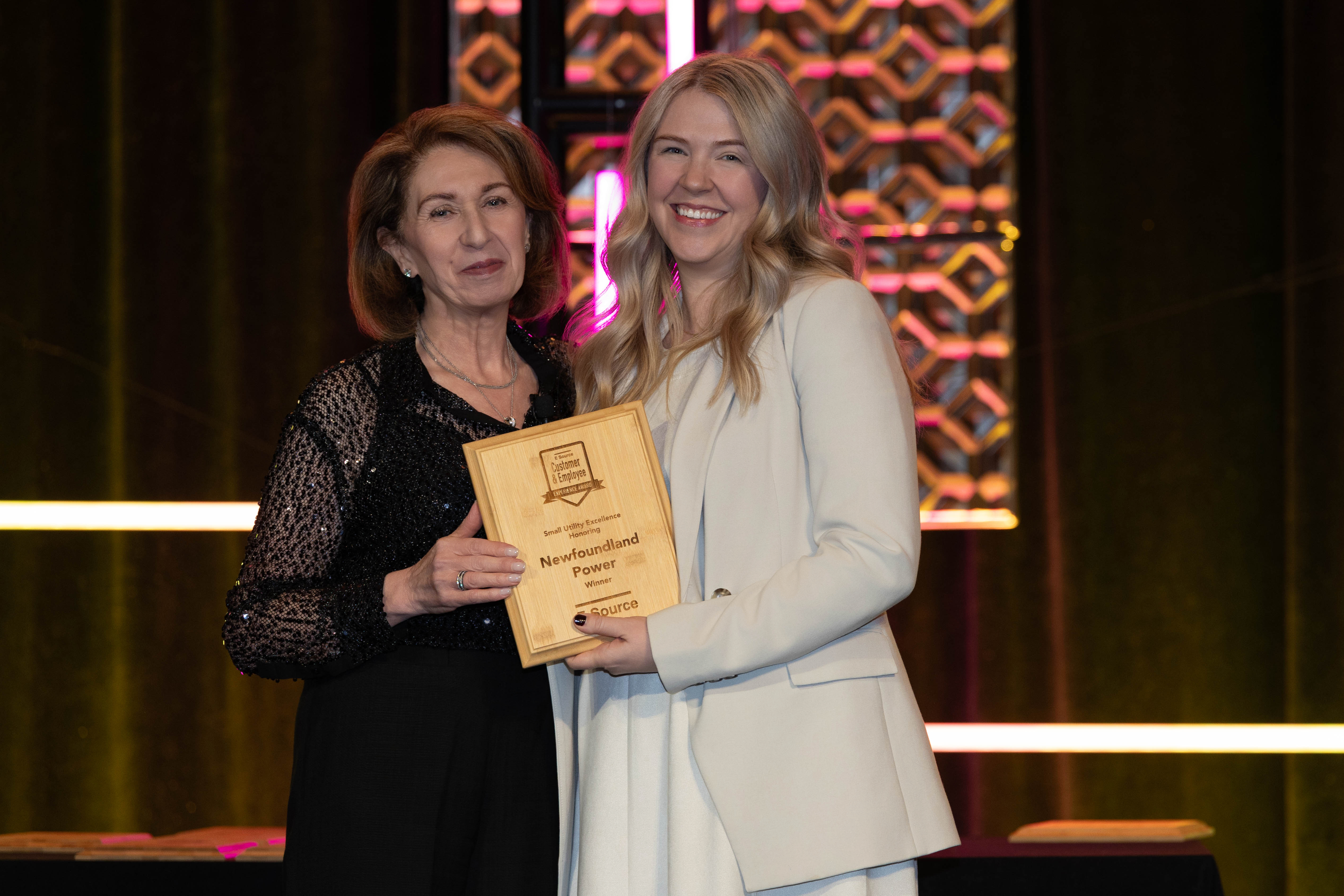 Two women stand on stage holding a wooden E Source award plaque together. The plaque reads Small Utility Excellence, Newfoundland Power, Winner. One woman wears a black sequined top. The other, smiling, wears a cream blazer. Pink-lit decorative panels glow behind them.