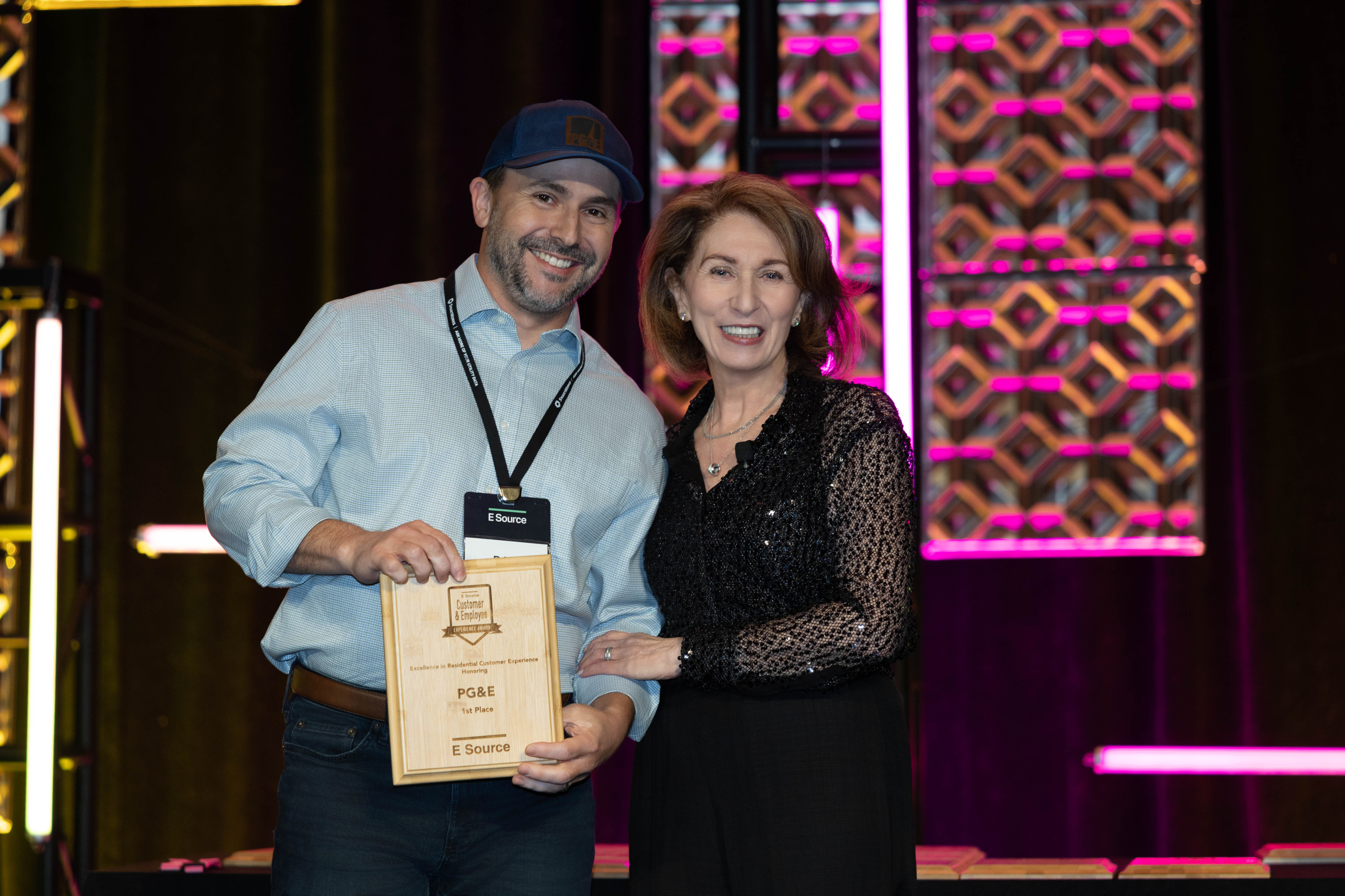 A smiling man in a baseball cap and light blue button-down shirt holds a wooden E Source award plaque reading PG&E, 1st Place. He stands on stage beside a smiling woman in a black sequined top, with pink-lit decorative panels behind them.
