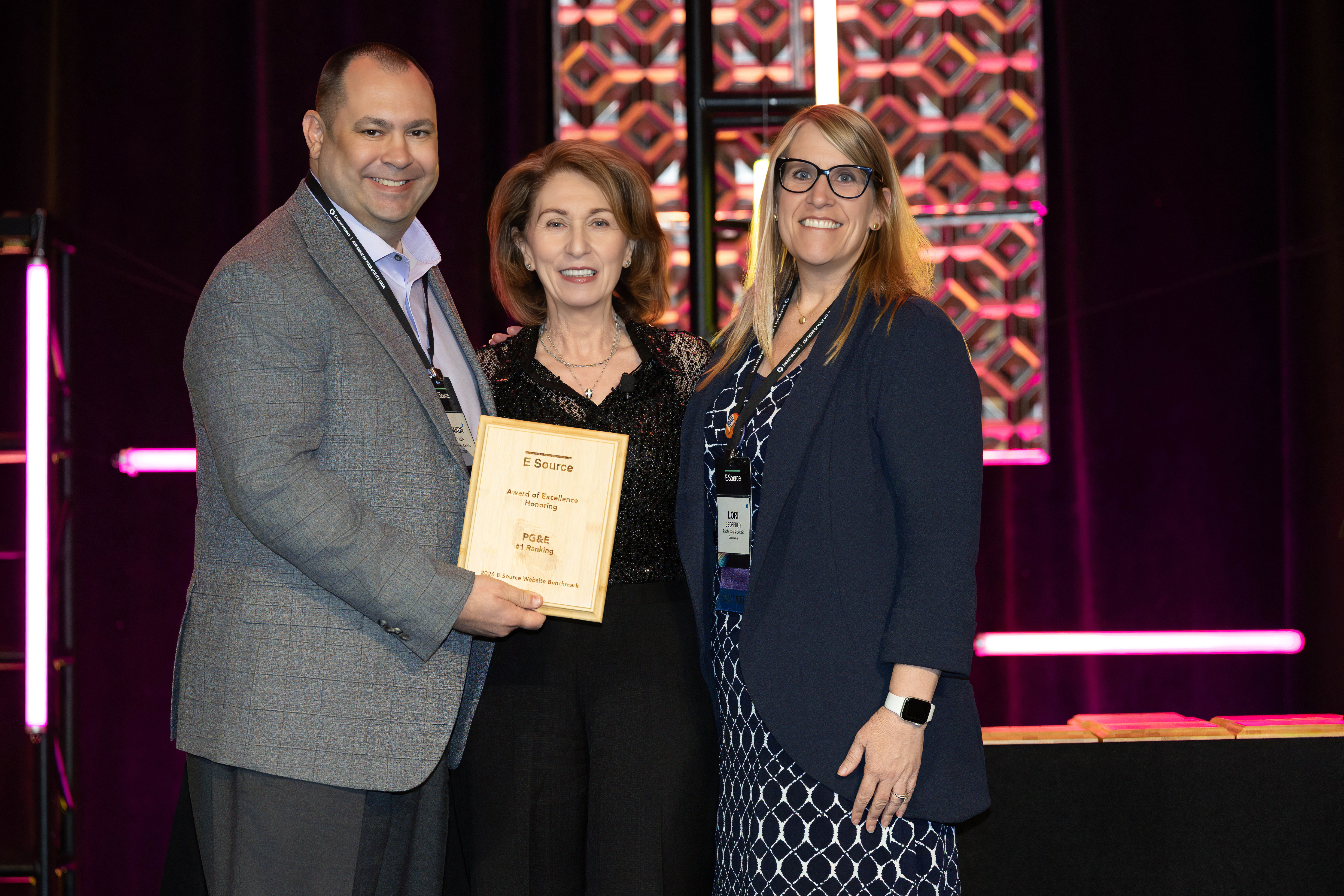 A man and two women stand on a stage. The man holds a wooden E Source award plaque reading Award of Excellence, PG&E, #1 Ranking. A woman in a black sequined top stands in the middle, and a woman in glasses and a navy blazer stands on the right. Pink-lit decorative panels glow behind them.