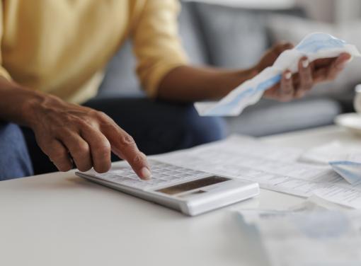Person using a calculator while holding receipts, with papers and a cup on a table.