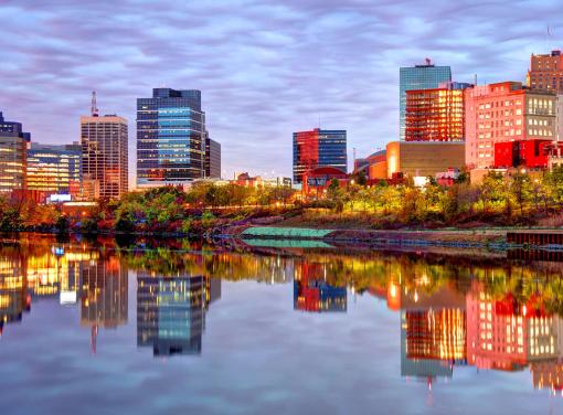 City skyline at sunset with colorful buildings reflected in a calm river under a cloudy sky.