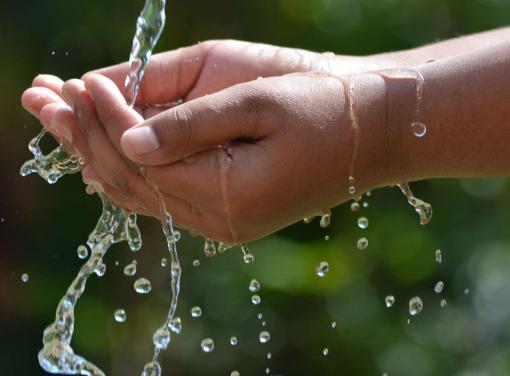 Close-up of hands cupping flowing water with droplets splashing against a blurred green background.