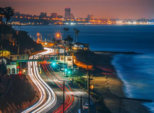 Nighttime coastal cityscape with illuminated roads showing light trails, a sandy beach, and distant city lights reflecting on calm water.