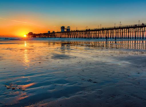 Sunset over a long pier extending into the ocean, with reflections on the wet sandy beach.
