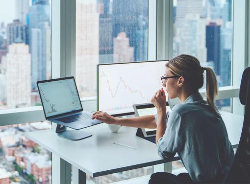 Person working at a desk with two monitors displaying financial charts in a high-rise office overlooking a city skyline.